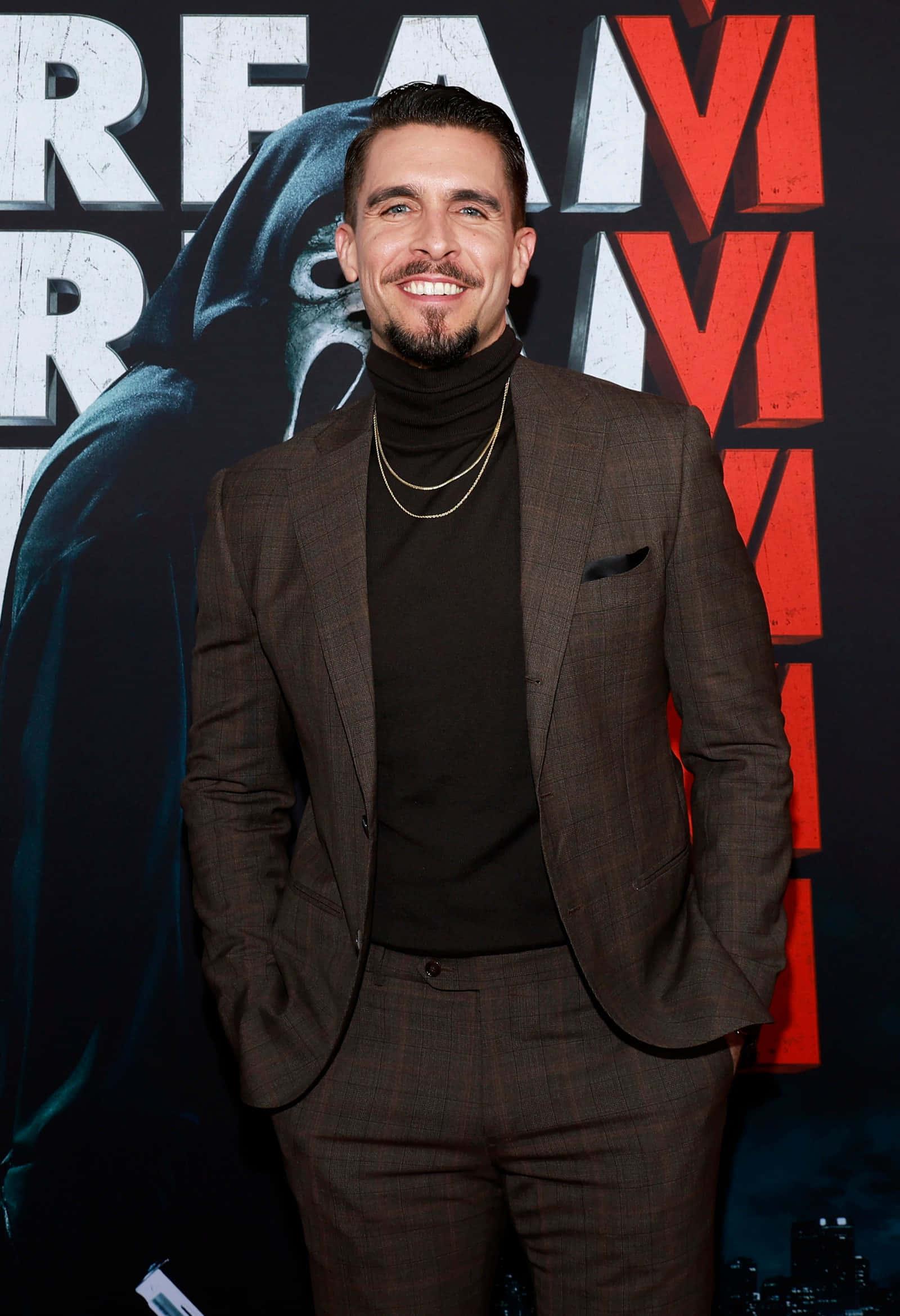 a man in a suit and a turtleneck standing in front of a poster — 1600x2340 wallpaper